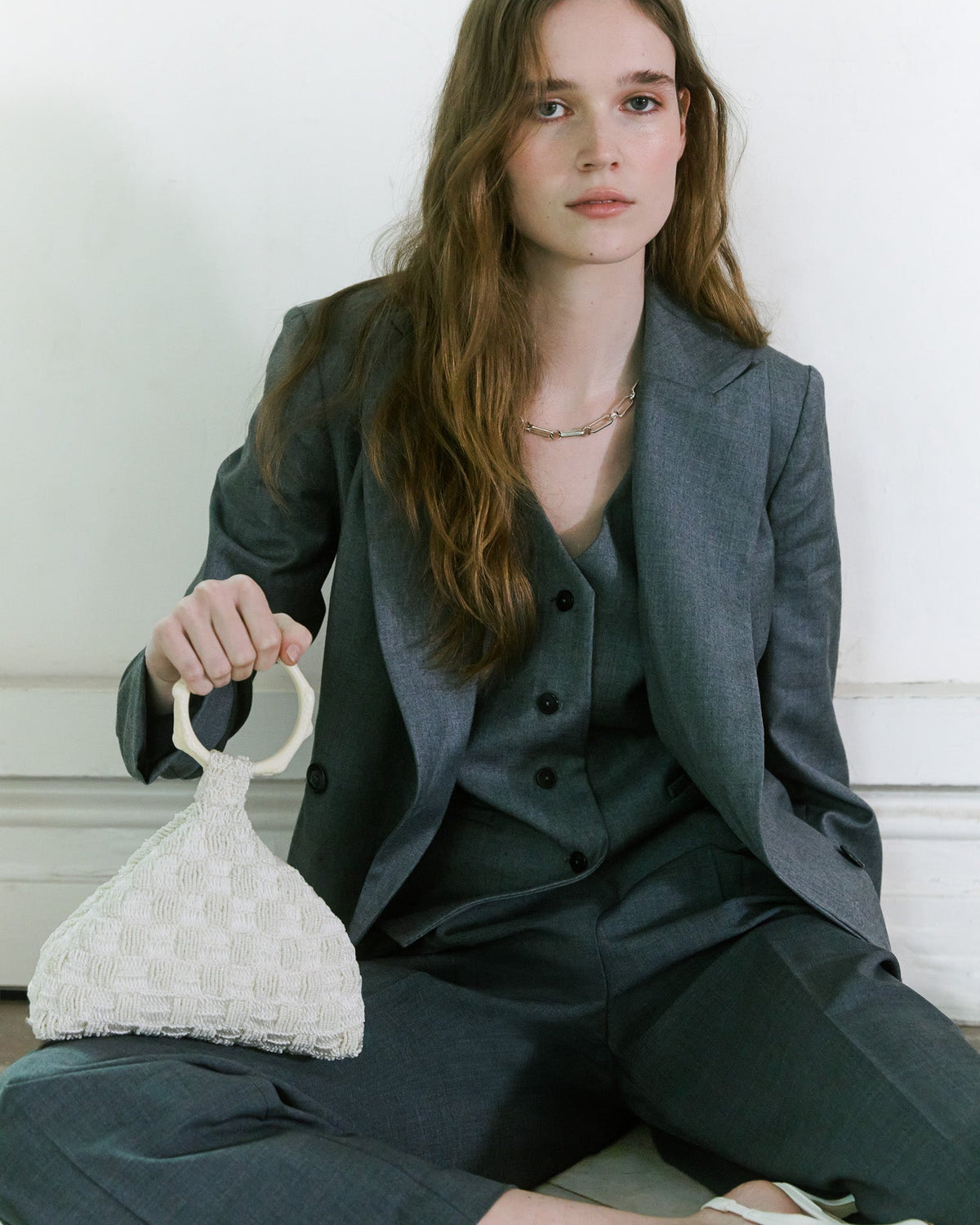 Model against a white wall, wearing a dark grey tailored suit and holding a small beaded and crochet off-white handbag with a circular handle by the hold