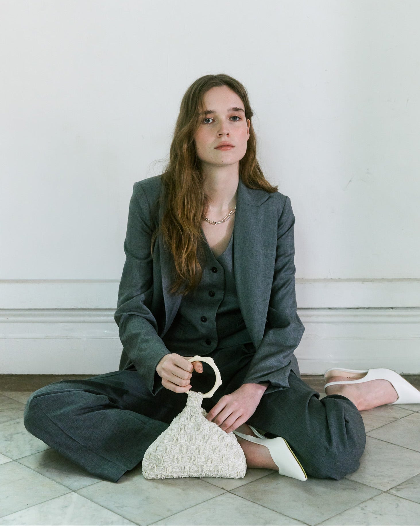 Model seated in charcoal tailoring carrying an ivory beaded and crochet handbag with sculptural handle by The Hold