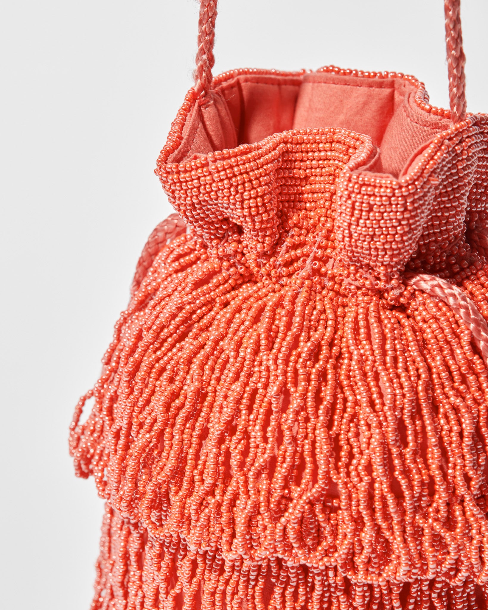 close up of detail of coral beaded bag showing beaded tassel fringe, a gathered drawstring top and long braided strap by the hold