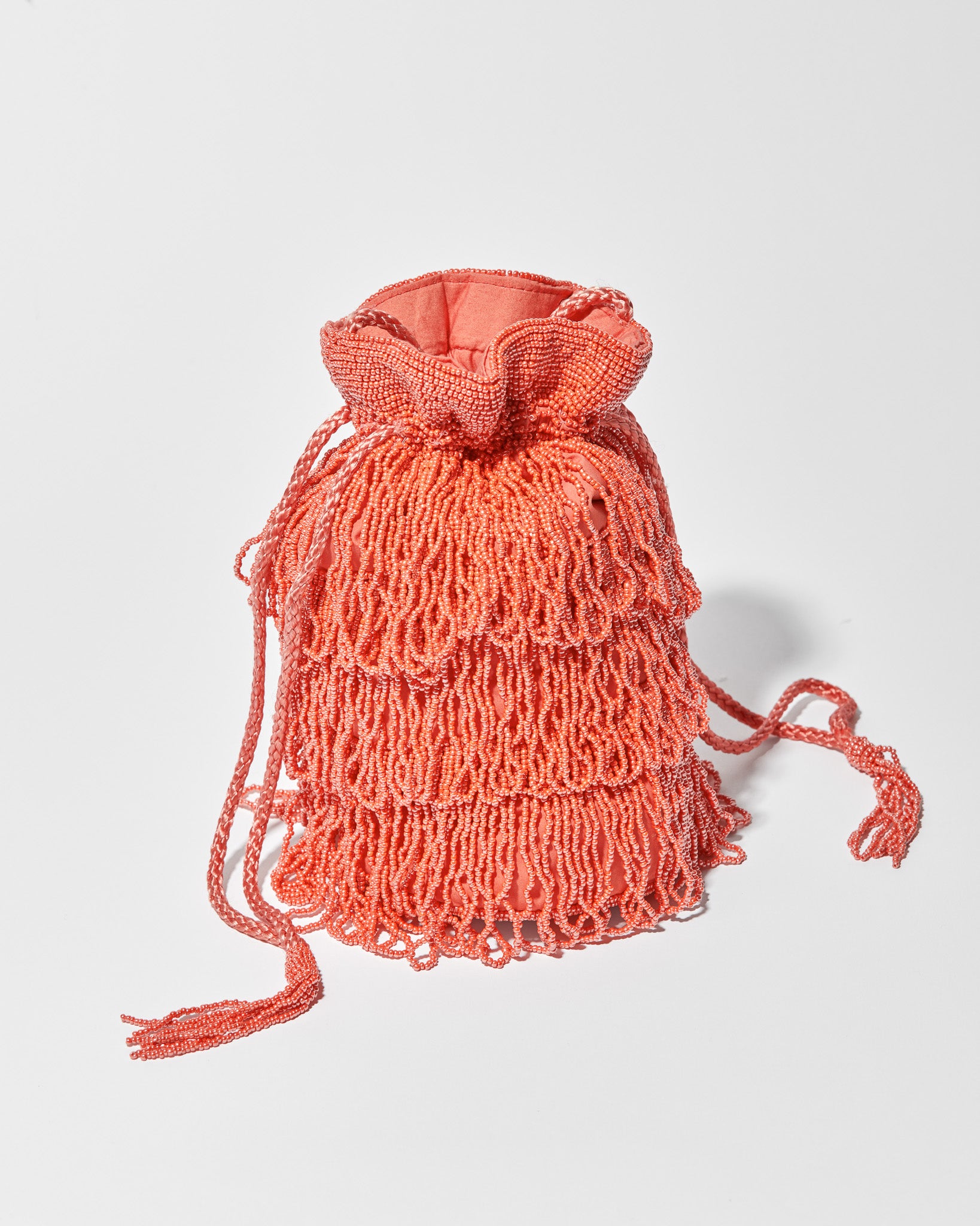 A coral beaded crossbody bag featuring three layers of beaded tassel fringe, a gathered drawstring top and long braided strap with beaded tassel ends sitting in studio
