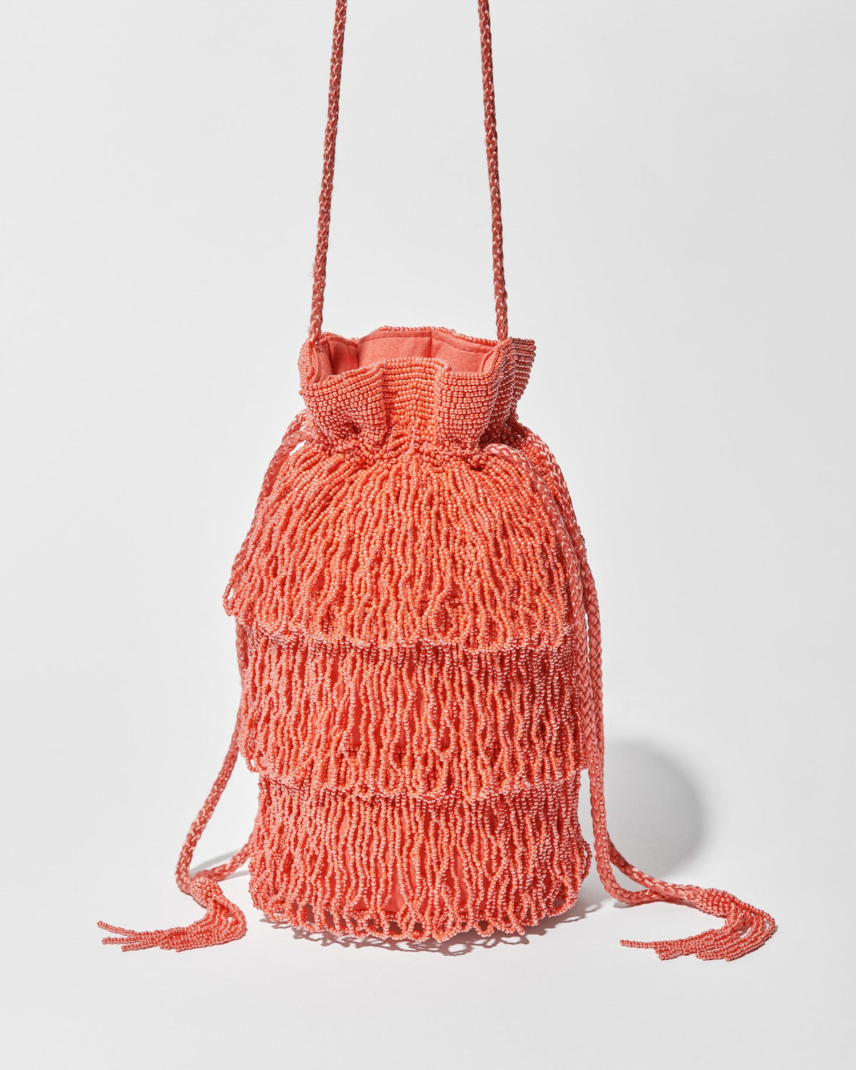A coral beaded crossbody bag featuring three layers of beaded tassel fringe, a gathered drawstring top and long braided strap with beaded tassel ends.
