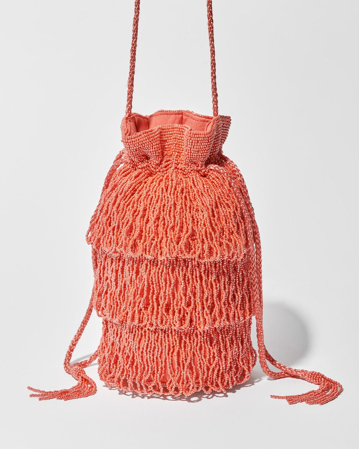 A coral beaded crossbody bag featuring three layers of beaded tassel fringe, a gathered drawstring top and long braided strap with beaded tassel ends.