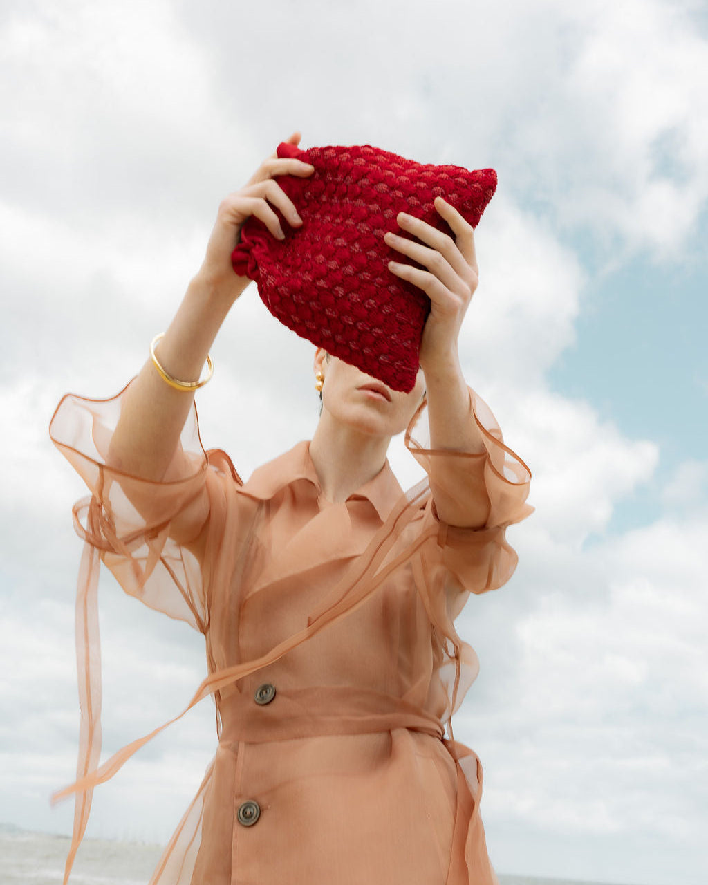 Model in a sheer peach dress holding the red beaded pouch against a beach backdrop, showcasing its tactile and expressive design.