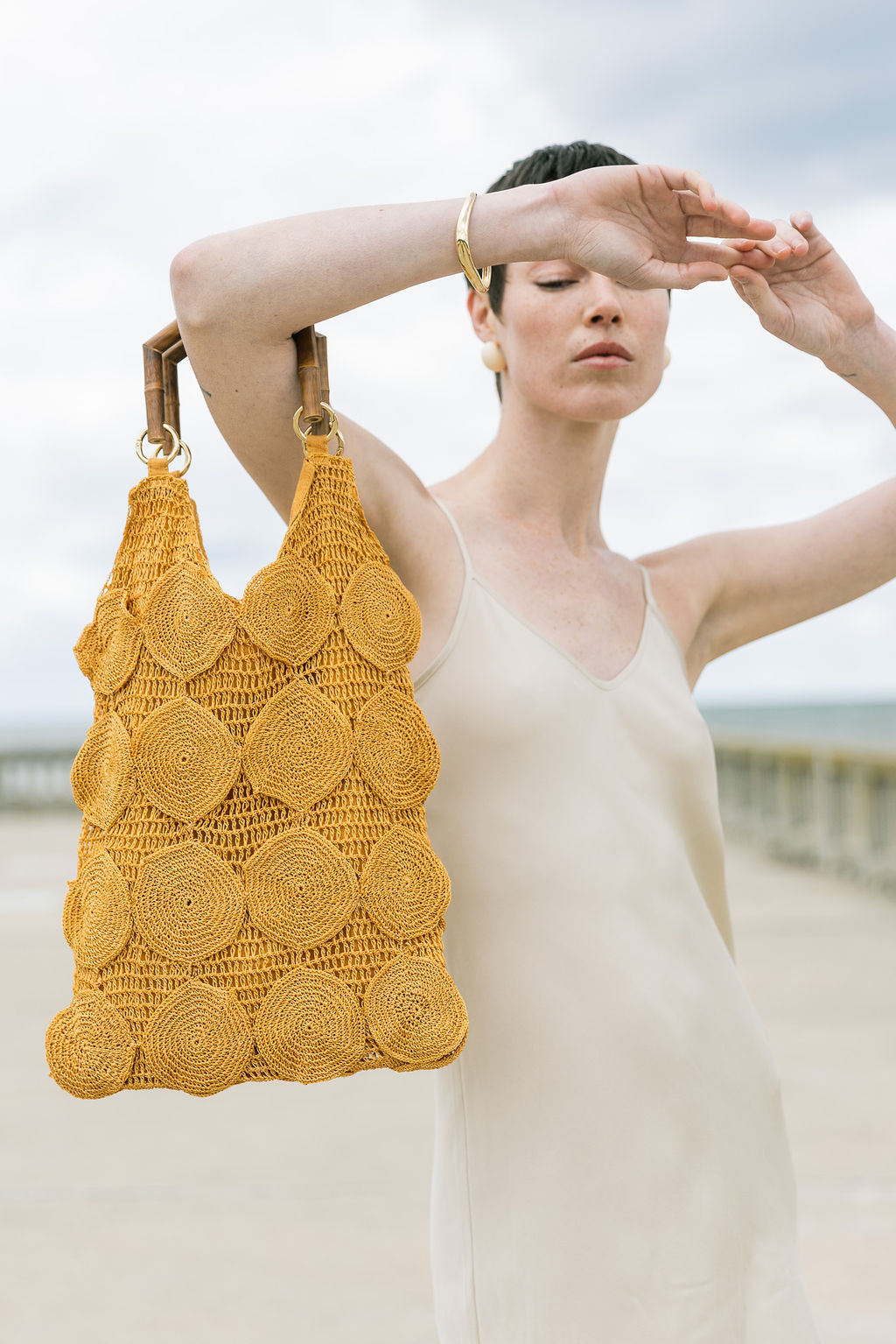 Model at the beach holding the ochre Enveloped Tote by its bamboo handles, showcasing its sculptural silhouette and artisanal texture.