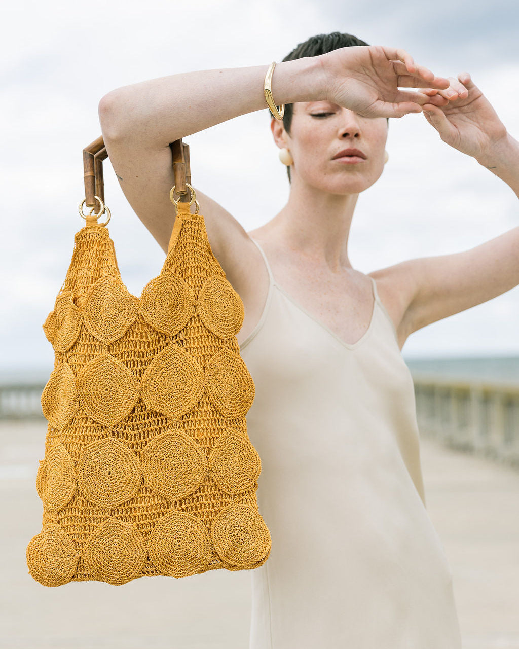 Model at the beach holding the ochre Enveloped Tote by its bamboo handles, showcasing its sculptural silhouette and artisanal texture.