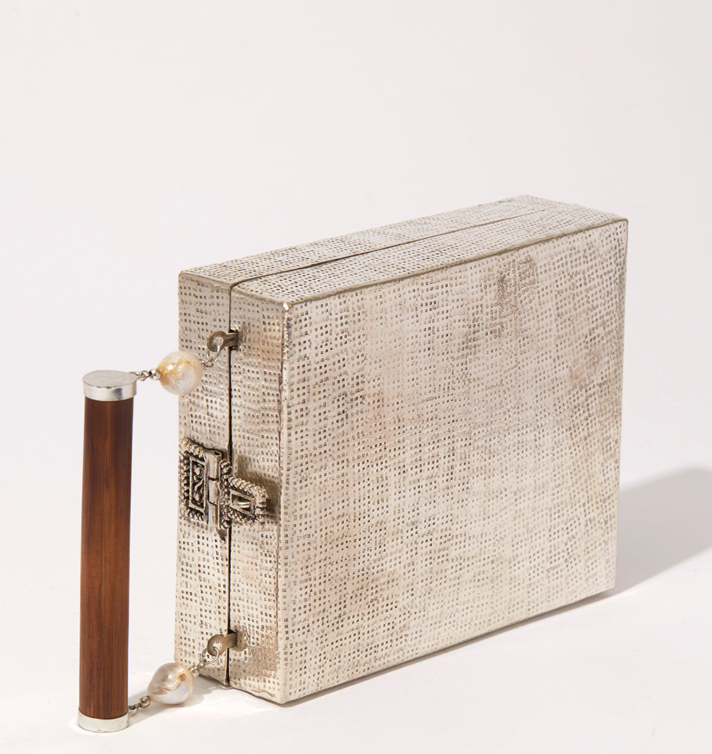 Three-quarter studio view of the Modern World brass bag featuring a silver hand-beaten texture and a wooden bar handle with pearl details.