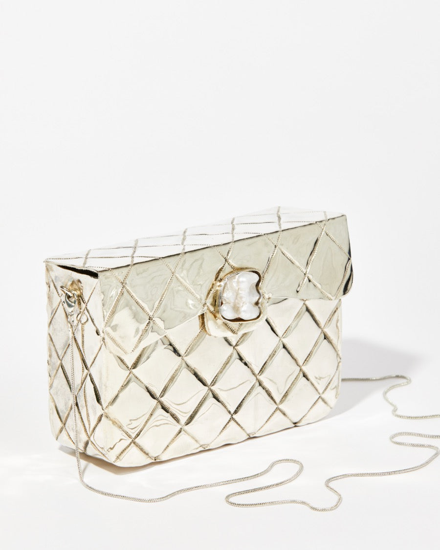 Angled view of the silver metal bag highlighting the detailed shell clasp with a Baroque pearl feature and the removable gold chain strap.