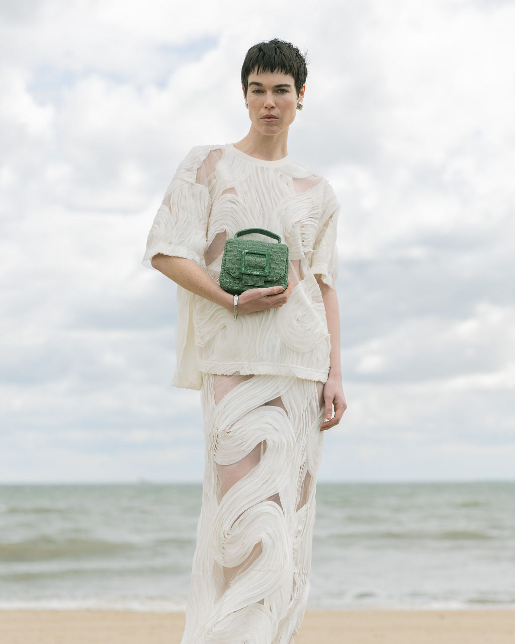 Model standing on a beach in a white lace dress holding the jade green beaded handbag, styled for an outdoor wedding or event.