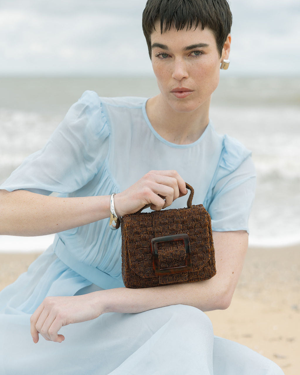 Model seated at the beach holding the rich brown Uncovered Layers bag, highlighting the warmth of the hand-dyed glass beads.