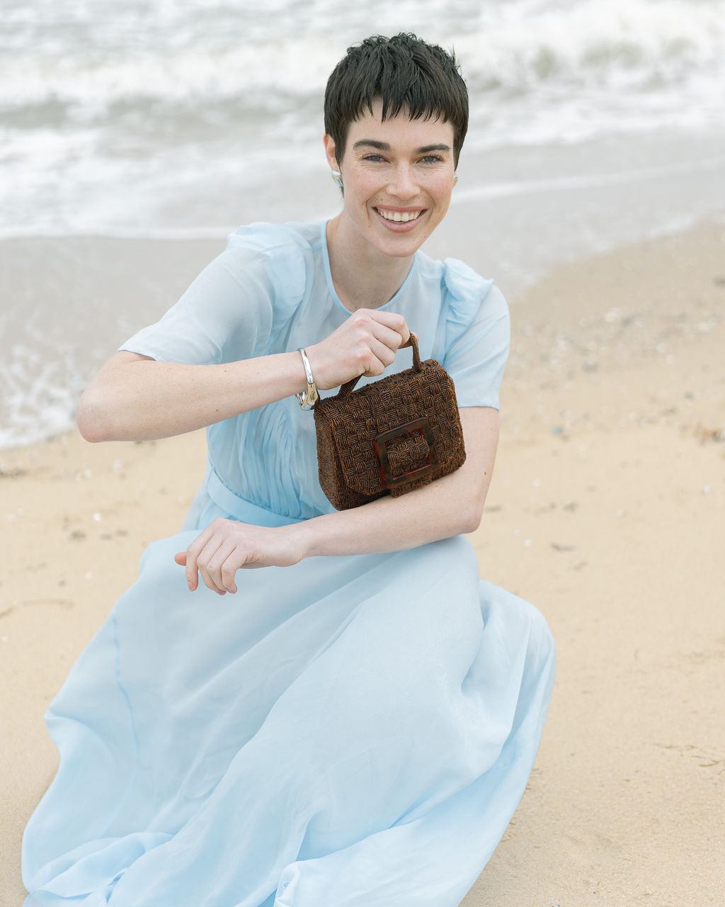Model in a light blue dress sitting on the sand with the brown Uncovered Layers bag, styled for an elegant daytime event or lunch.