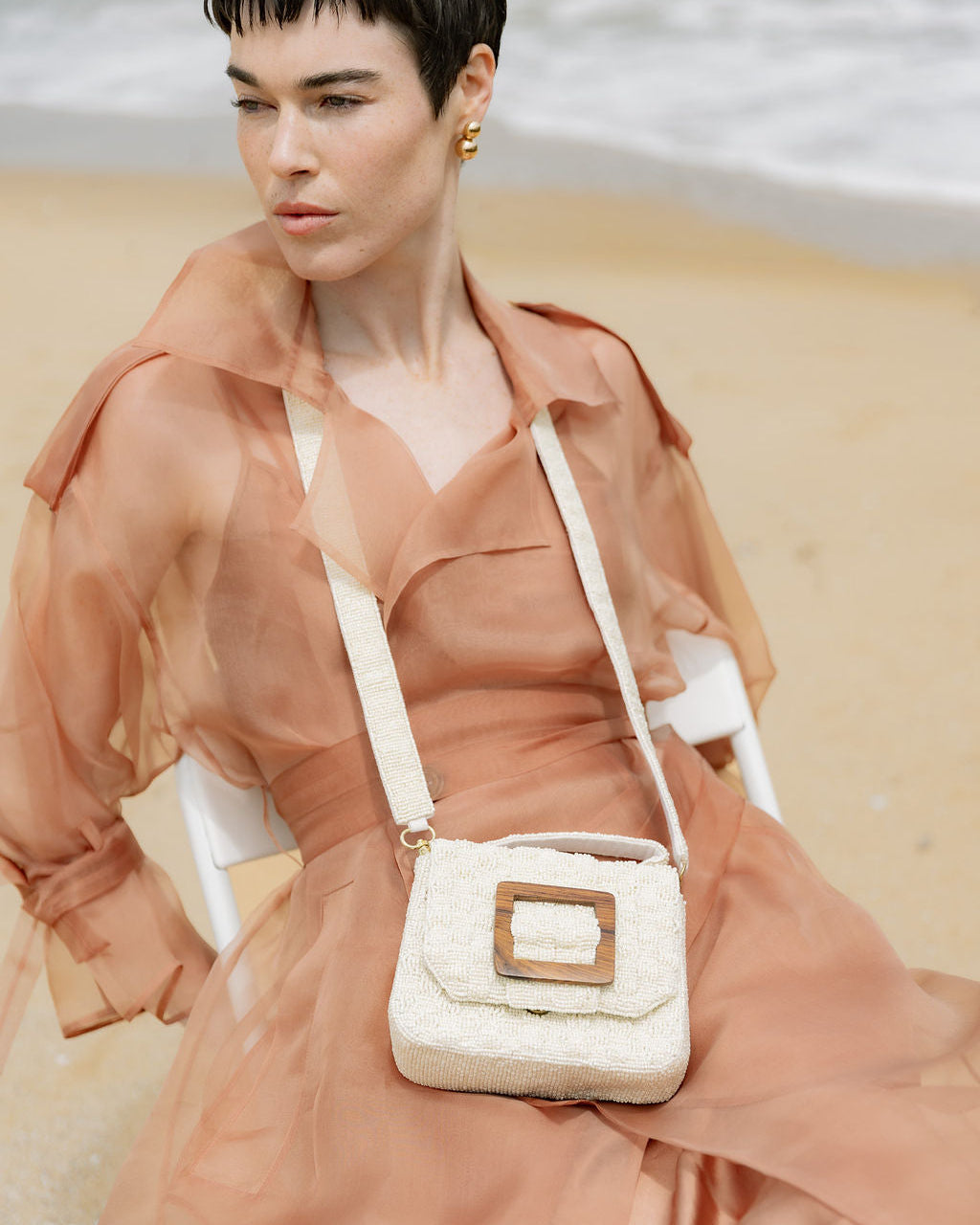 Model seated on a beach in a sheer peach dress, holding the white beaded Uncovered Layers bag as a trendy accessory for outdoor events.