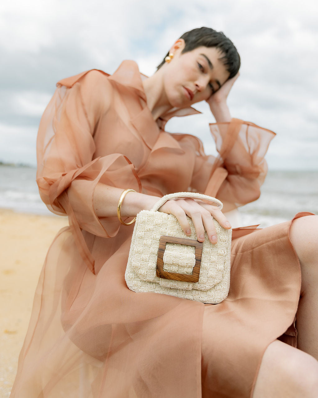 Model in a sheer peach dress holding the white beaded bag against a coastal backdrop, showcasing its soft luminosity and sculptural shape.