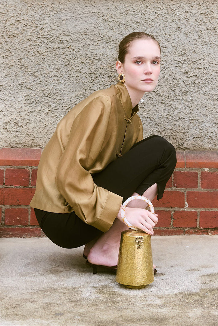 Model holding a gold metallic top-handle bag with marble-effect resin handle, styled with a bronze blouse and black trousers.