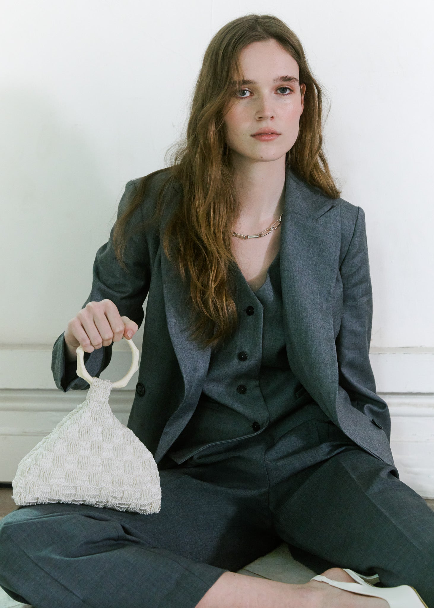 Model against a white wall, wearing a dark grey tailored suit and holding a small beaded and crochet off-white handbag with a circular handle by the hold