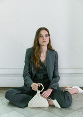 Model seated in charcoal tailoring carrying an ivory beaded and crochet handbag with sculptural handle by The Hold