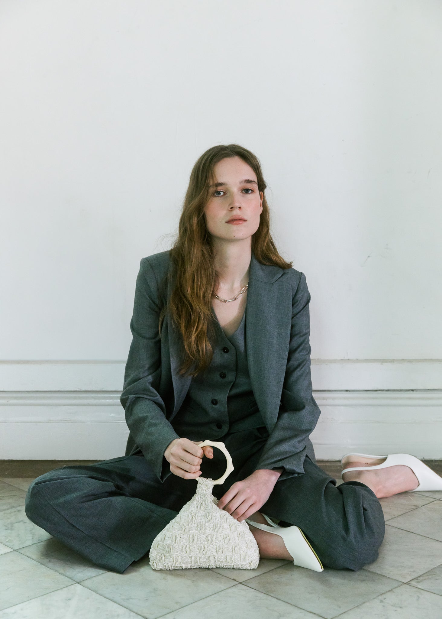 Model seated in charcoal tailoring carrying an ivory beaded and crochet handbag with sculptural handle by The Hold