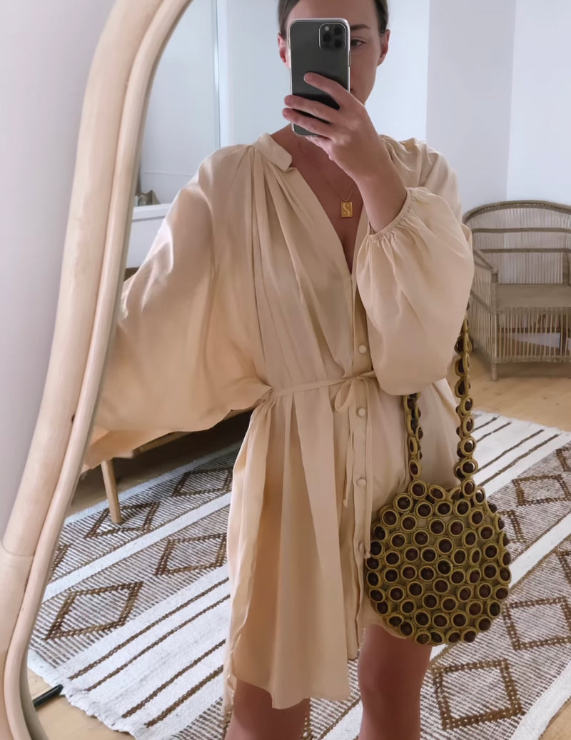 A woman , wearing a beige long-sleeve mini dress  carrying a wooden beaded shoulder bag shown how it can sit on your arm as well.