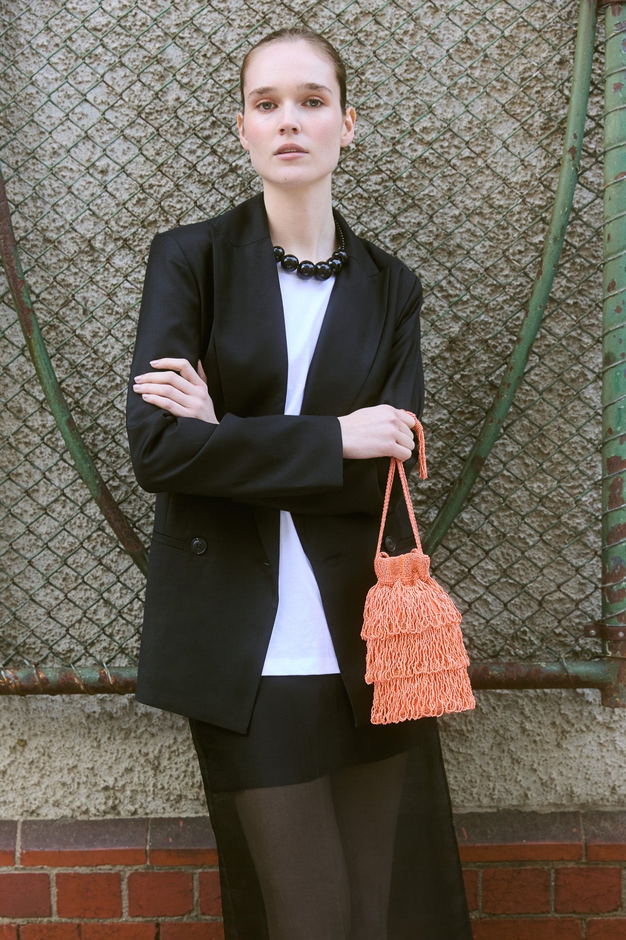 Model holding a coral beaded fringe crossbody bag with layered tassels, styled with a black blazer and sheer skirt