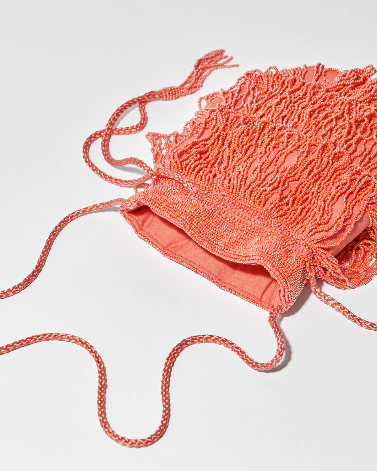 Coral beaded bag laid flat, featuring three layers of tassel fringe, a gathered drawstring top, and a long braided strap finished with beaded tassel ends.