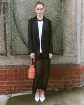 A woman stands outdoors awearing a black  blazer holding a coral beaded tassle handbag.