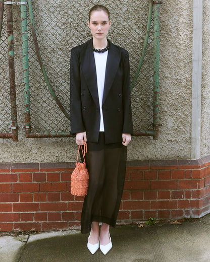 A woman stands outdoors awearing a black  blazer holding a coral beaded tassle handbag.