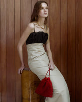 A woman wearing a black and beige  dress leans against a wooden barrel in, holding a red beaded drawstring bag.
