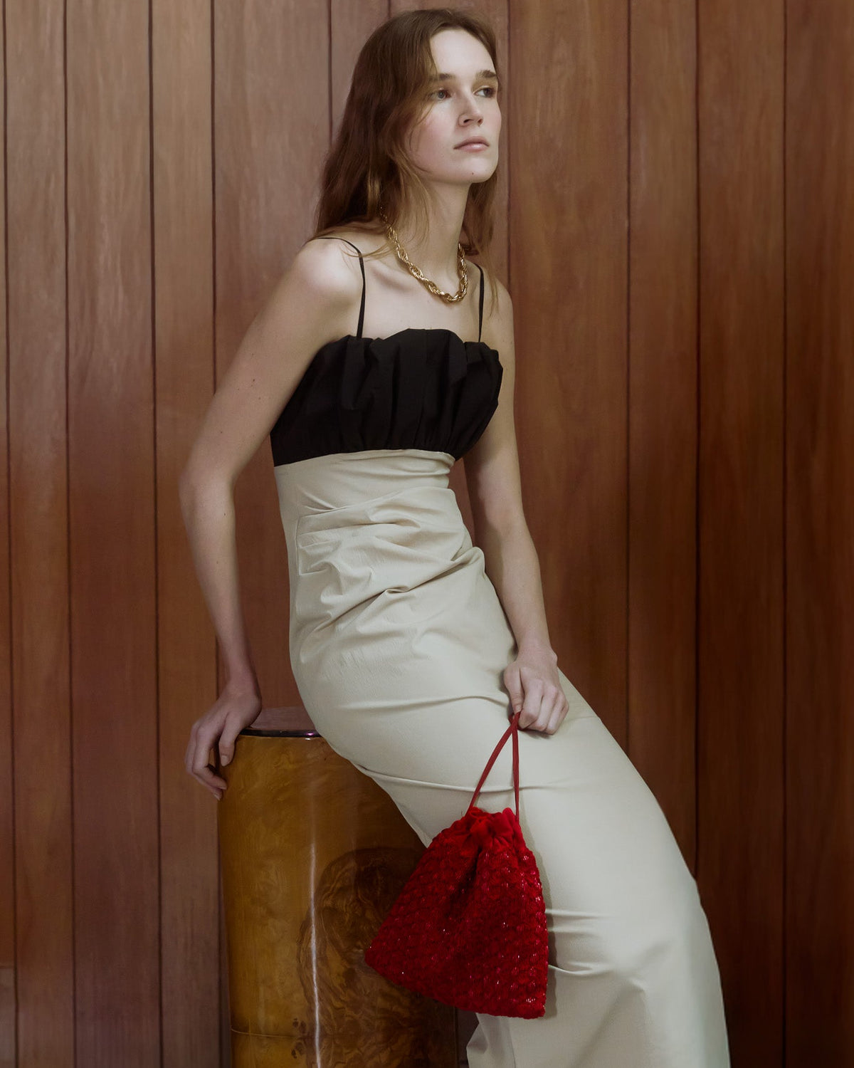 A woman wearing a black and beige  dress leans against a wooden barrel in, holding a red beaded drawstring bag.