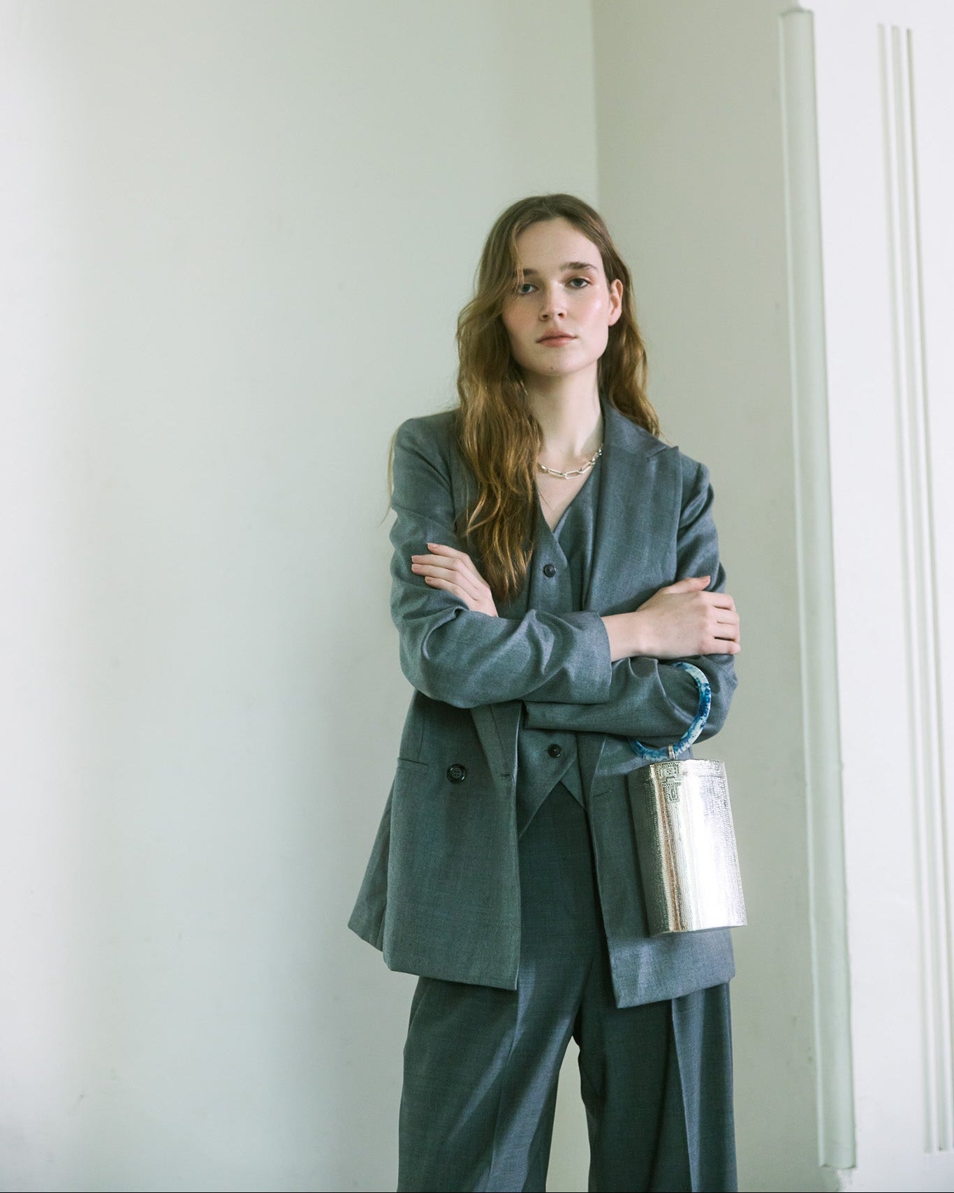 Model in charcoal suit wearing silver metal bucket handbag on her arm with a sculptural blue handle by The Hold.