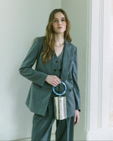Model in charcoal suit holding a silver metal bucket handbag with a sculptural blue handle by The Hold.