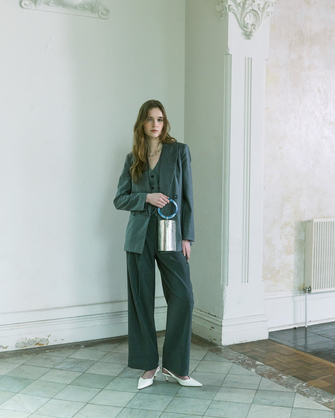Model in charcoal tailoring holding a silver metallic top handle handbag with a sculptural blue handle by The Hold.