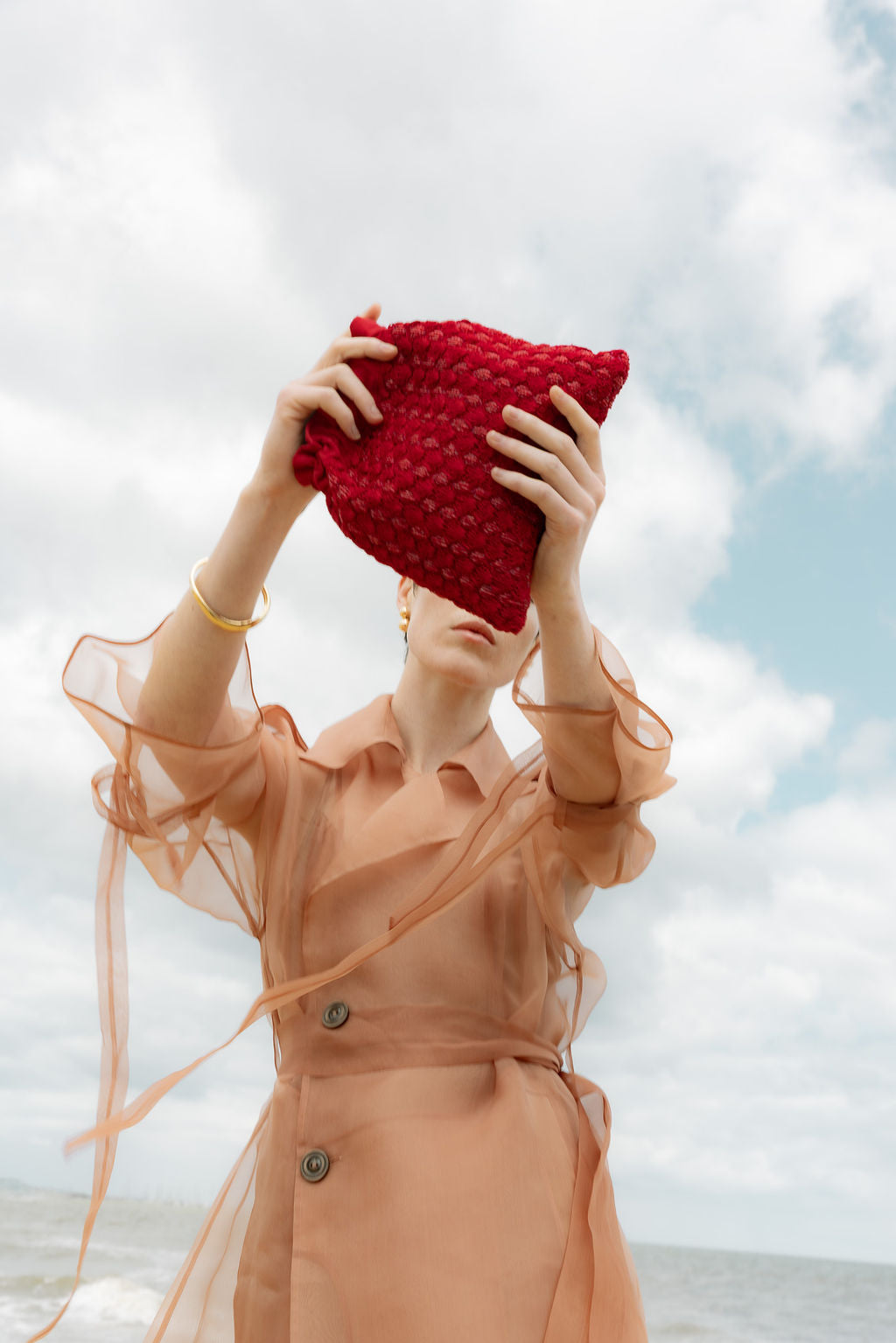 Model in a sheer peach dress holding the red beaded pouch against a beach backdrop, showcasing its tactile and expressive design.