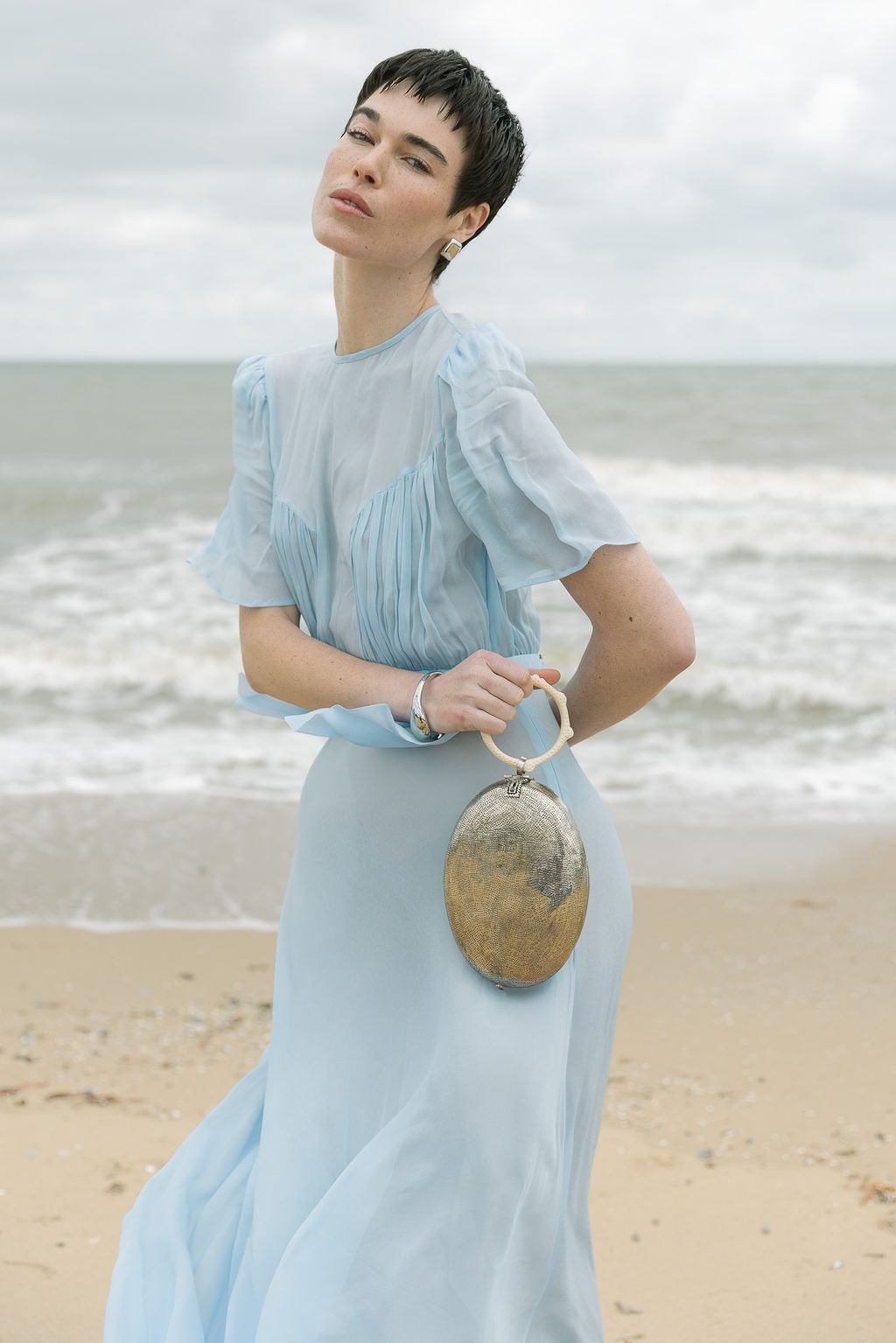 Model in a pale blue dress holding the silver sculptural metal bag by its resin handle against a coastal beach backdrop.