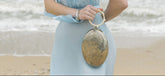 Model in a pale blue dress holding the silver sculptural metal bag by its resin handle against a coastal beach backdrop.
