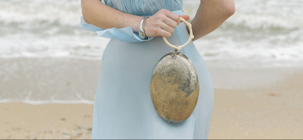 Model in a pale blue dress holding the silver sculptural metal bag by its resin handle against a coastal beach backdrop.
