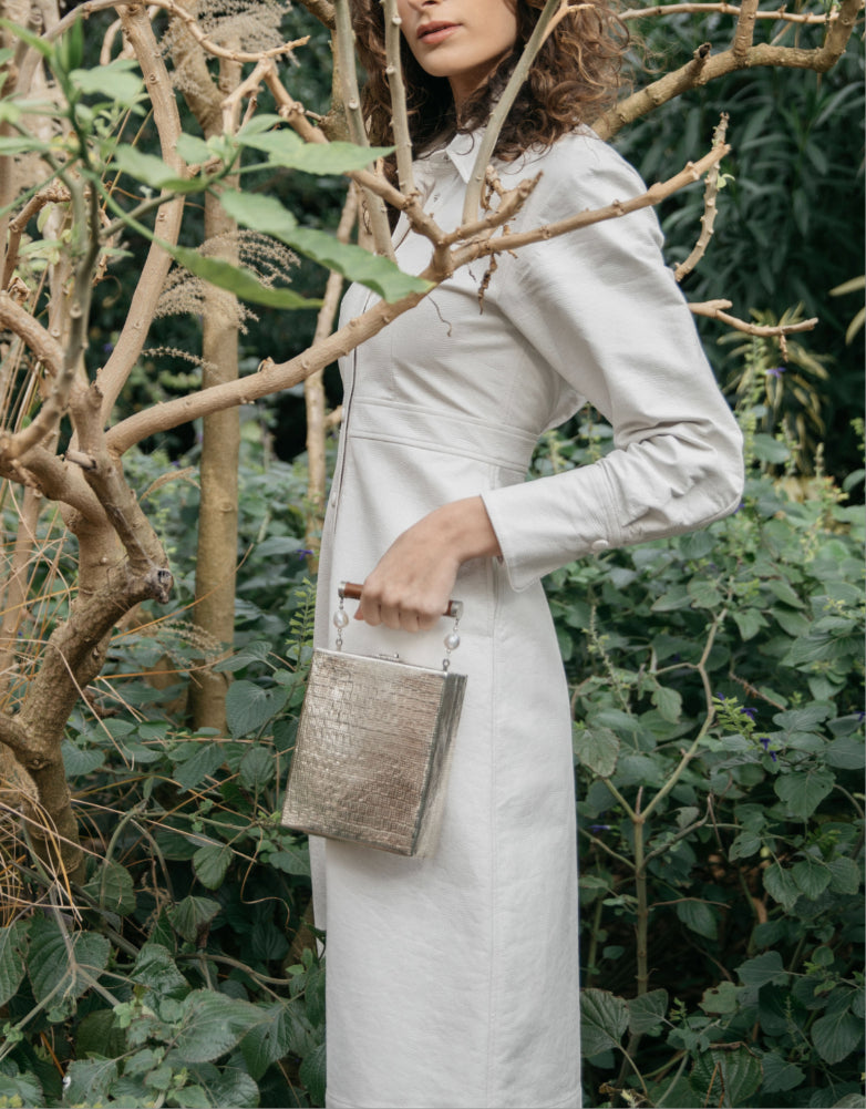 Model carrying the Modern World silver metal bag in a lush garden setting, demonstrating its elegant 15cm height and sculptural form.