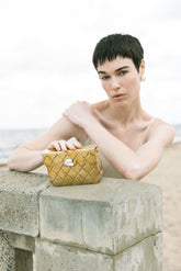Model holding the gold Our Longing metal bag against a coastal backdrop, showcasing its reflective hand-beaten brass surface.