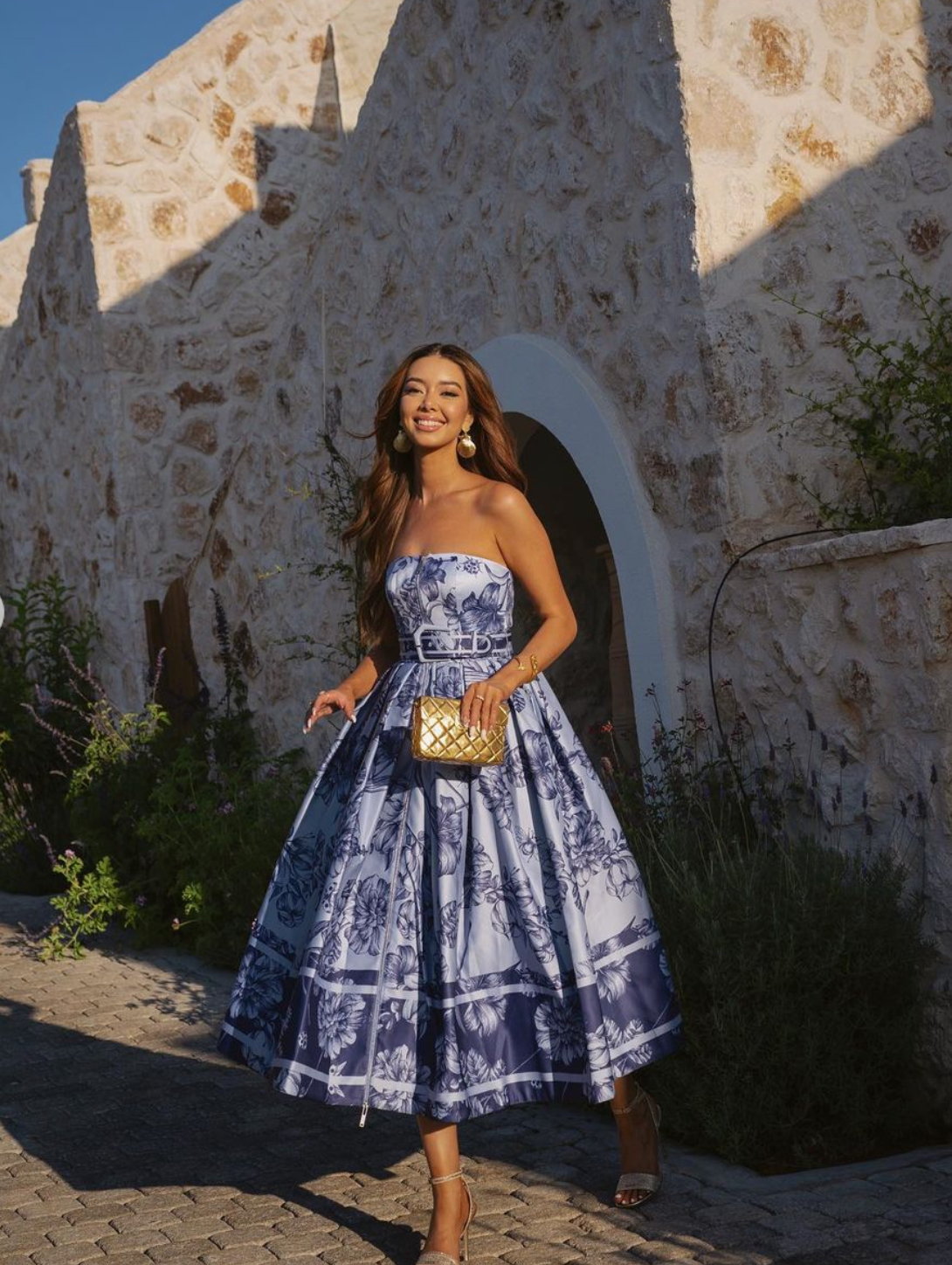 Model in a blue floral gown carrying the gold Our Longing bag, showcasing its presence as a striking event accessory.