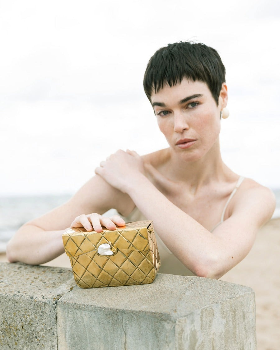 Model holding the gold Our Longing metal bag against a coastal backdrop, showcasing its reflective hand-beaten brass surface.