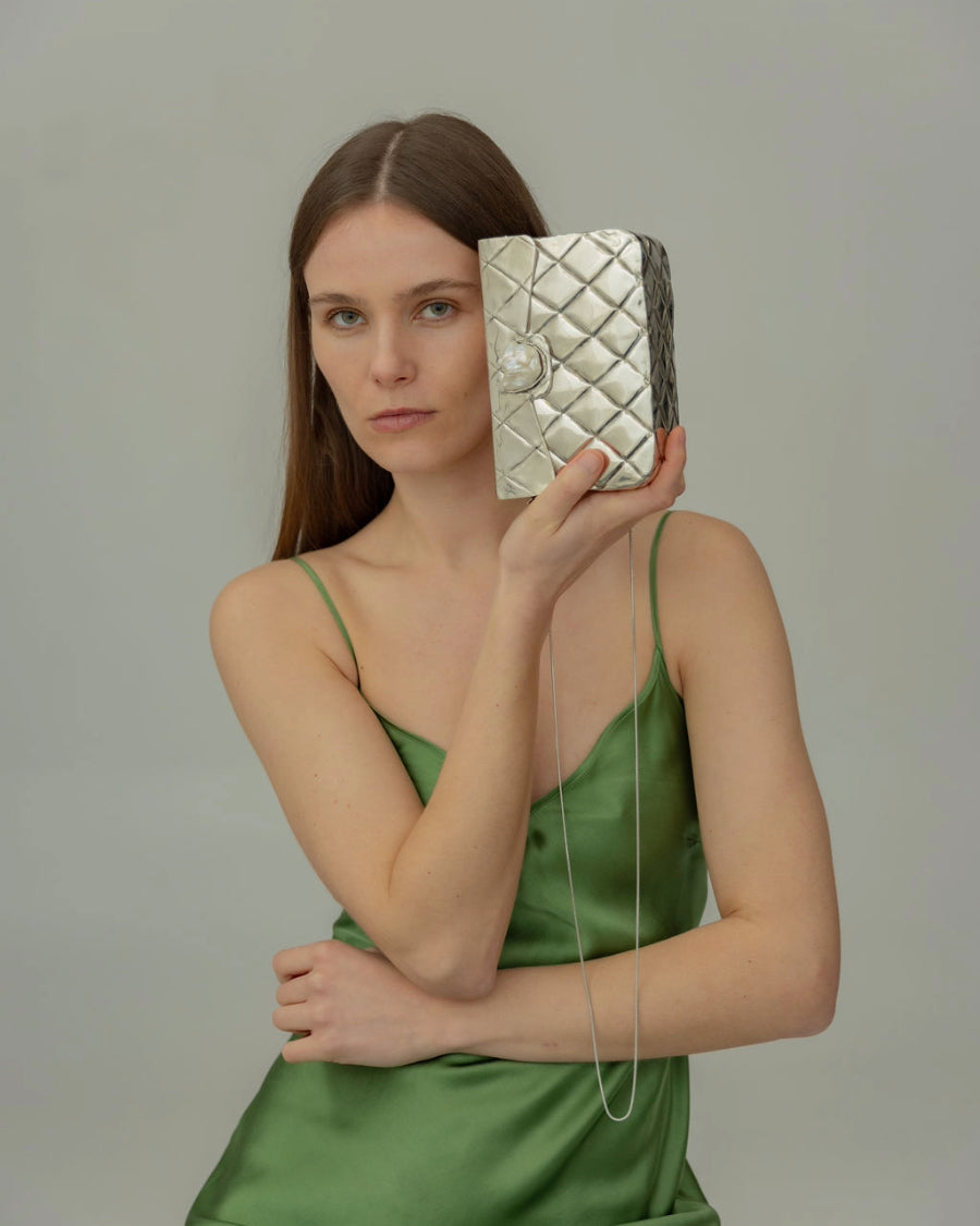 Model in a green slip dress holding the silver Our Longing clutch, showcasing its reflective hand-beaten surface and elegant scale.
