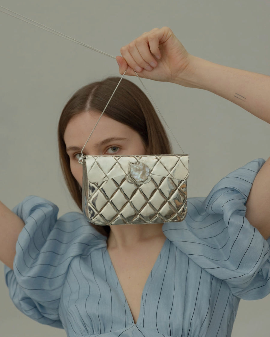 Model in a blue puff-sleeve dress holding the silver Our Longing bag, emphasising its presence as a piece of wearable art for events.