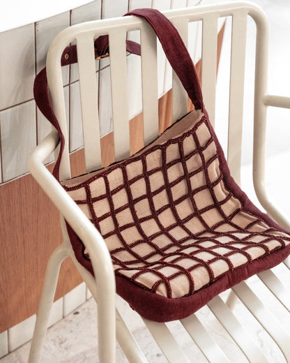The Returning Form beaded tote draped over a modern white chair, showcasing its sculptural grid pattern and hand-dyed glass beads.