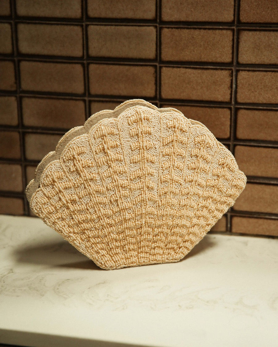 Artisan-made shell-shaped beaded clutch in champagne resting on a table, highlighting the intricate hand-dyed glass beads.