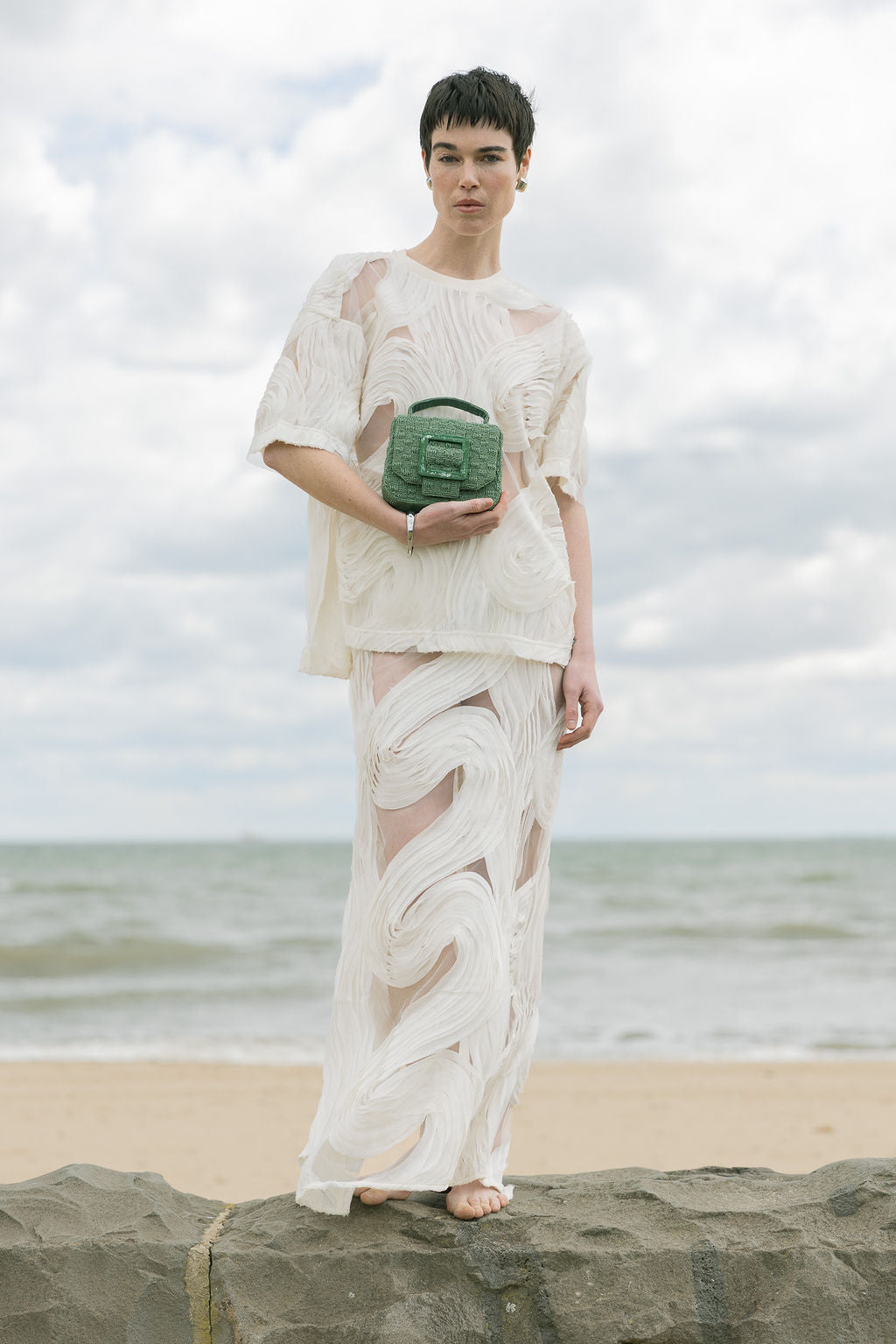 Model standing on a beach in a white lace dress holding the jade green beaded handbag, styled for an outdoor wedding or event.