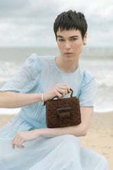 Model seated at the beach holding the rich brown Uncovered Layers bag, highlighting the warmth of the hand-dyed glass beads.