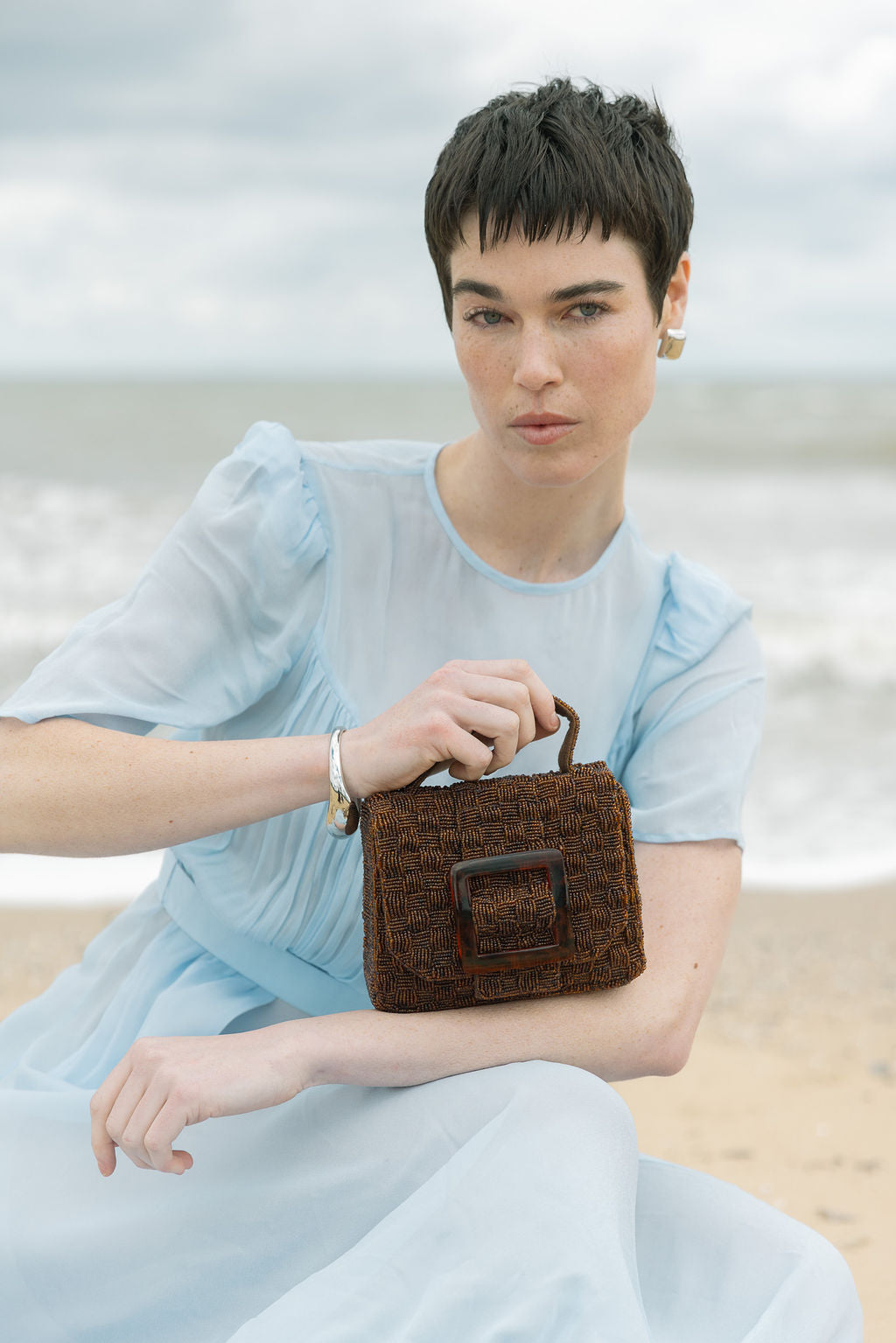 Model seated at the beach holding the rich brown Uncovered Layers bag, highlighting the warmth of the hand-dyed glass beads.