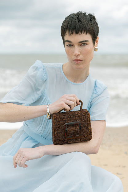 Model seated at the beach holding the rich brown Uncovered Layers bag, highlighting the warmth of the hand-dyed glass beads.