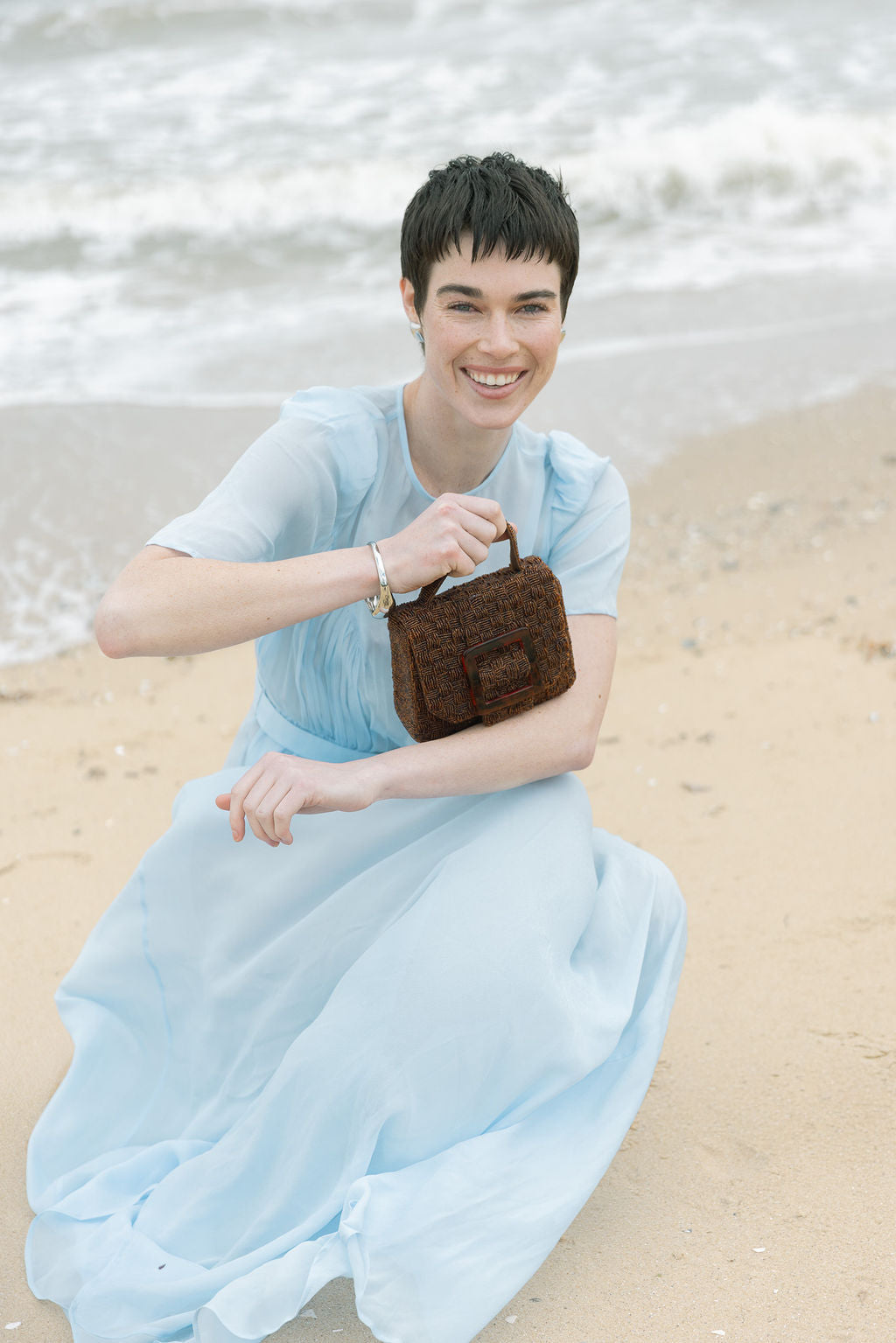 Model in a light blue dress sitting on the sand with the brown Uncovered Layers bag, styled for an elegant daytime event or lunch.