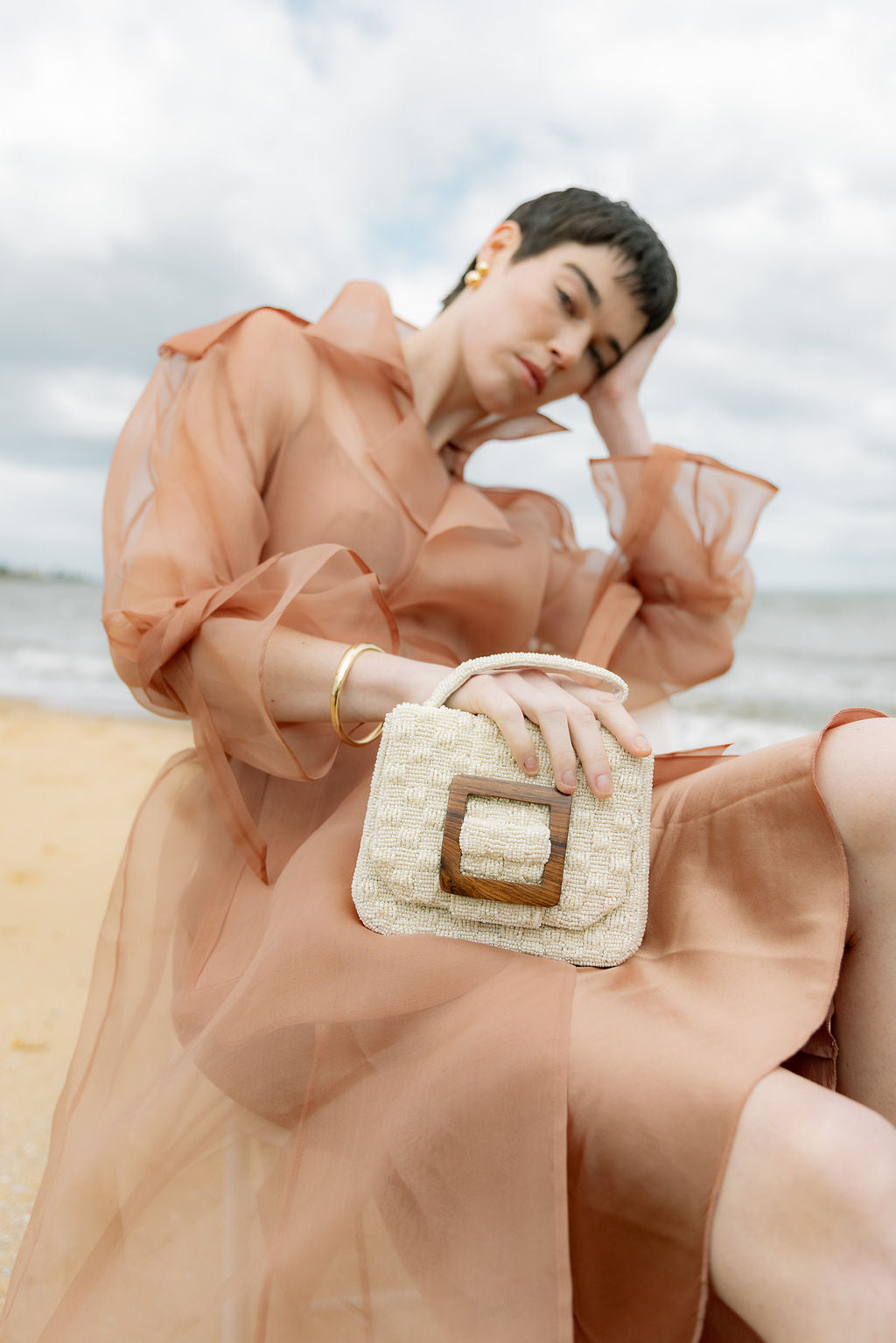 Model in a sheer peach dress holding the white beaded bag against a coastal backdrop, showcasing its soft luminosity and sculptural shape.