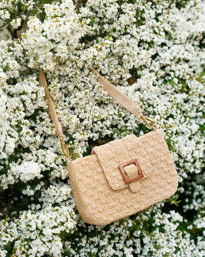 Lifestyle shot of the tan Familiar Way beaded bag hanging amidst white flowers, highlighting its natural, artisanal aesthetic.