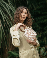 Model in a cream jumpsuit holding the Symbolic Path beaded bag in a lush green garden setting, showcasing its elegant handheld style.